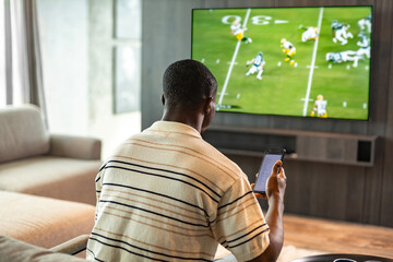 African man holding smartphone with sports bets app while watching American football match at home in living room.