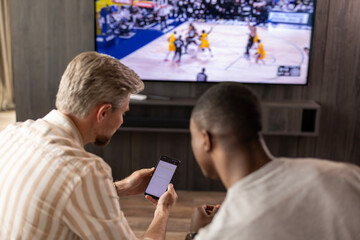 Two friends holding smartphone with sports bets app while watching basketball match at home in living room.