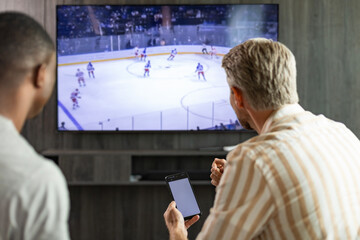 Two friends holding smartphone with sports bets app while watching ice hockey match at home in living room.