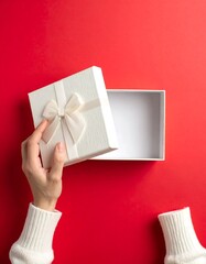 A close-up view of a person's hand opening an empty rectangular gift box tied with a satin ribbon on a vibrant red backdrop