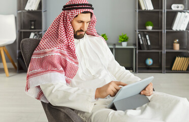 Arab businessman in office using tablet on chair. Wearing a keffiyeh and thobe, he is browsing data, reviewing mail, and staying focused in a tidy lounge in modern office. Corporate workflow concept.