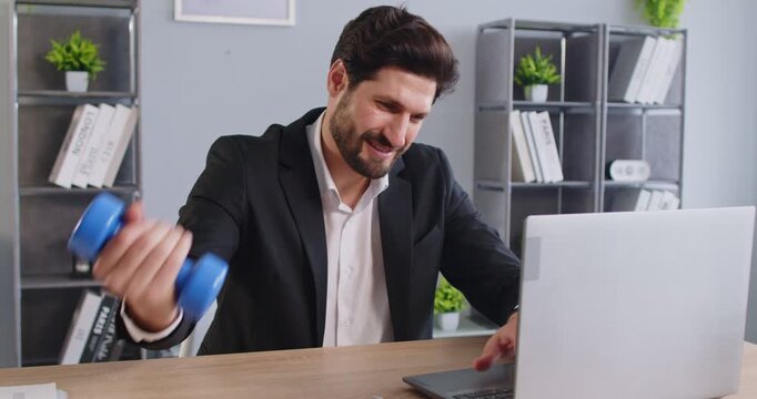 Businessman sitting at desk, holding blue dumbbell, exercising his arm, looking at laptop screen, smiling, combining work and fitness, happy energized to be active, sporty, strong and fit in office