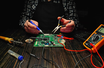 A person is focused on repairing a circuit board using various tools like a multimeter, pliers, and soldering iron on a dark wooden table at night