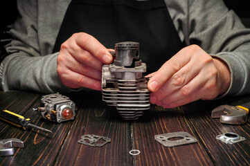 Hands focus on assembling a small trimmer engine part on a wooden table in a workshop. Various tools and components are spread out. This happens in the evening