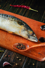 A whole fish lies on a wooden cutting board next to a spoon filled with black pepper. Various spices are scattered around, indicating preparation for cooking in a home kitchen during the evening
