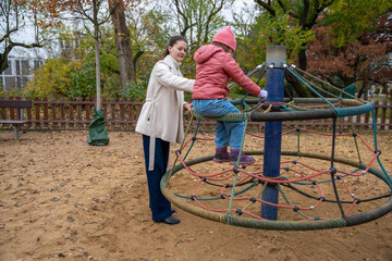 Mother Steadying Daughter On Spinning Roundabout Active Play With Supervision, Autumn Park, Jacket And Hat, Trusting Grip, Energetic Circle Of Motion