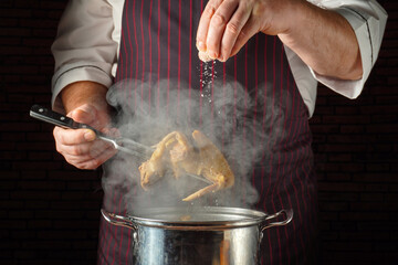 A chef holds a piece of chicken above a pot while sprinkling salt onto it. Steam rises from the pot, showing an active kitchen scene during cooking