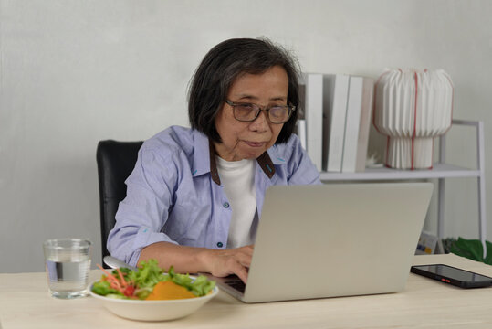 Senior Asian woman working on laptop and typing while eating healthy salad lunch at home. Elderly digital lifestyle, remote work, freelance, and multitasking concept.