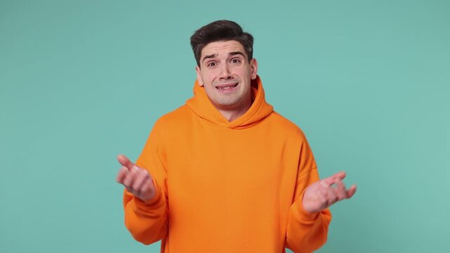 Hesitant indecisive young man he wearing orange hoodie feels doubtful spreading hands say oops ouch oh omg i am so sorry isolated on plain pastel light blue cyan background studio. Lifestyle concept