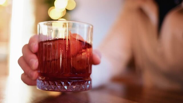 Close up of a hand holding a glass filled with a red cocktail, ice cubes, and citrus peel
