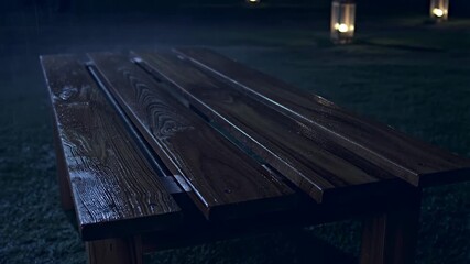 Wooden table with raindrops in a serene outdoor setting illuminated by lanterns. Ideal for cozy gatherings and outdoor dining experiences