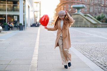 Woman walks on city street holding red heart balloon during sunset