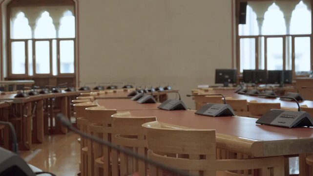 Empty conference room featuring wooden tables and professional microphones for business meetings. Rows of chairs in vacant boardroom of university or government office building environment.