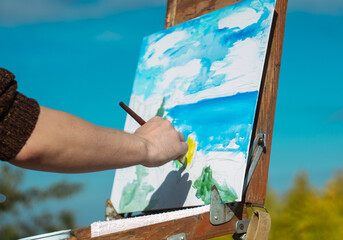 Close up of an artist's hand painting a landscape on a canvas in an outdoor setting with vibrant sky.