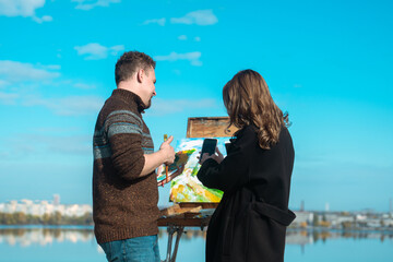 A man paints on an easel while a woman observes, both enjoying the sunny weather by a calm lake.