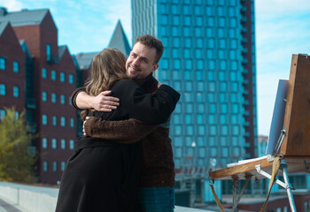 A couple hugs warmly on a rooftop with a cityscape background, featuring red brick buildings and an art easel.
