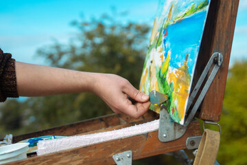 A close-up of an artist using a palette knife to paint a colorful landscape on an easel in an outdoor setting.