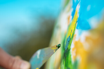 A close-up shot of an artist's hand using a palette knife to create a vibrant painting on canvas outdoors.