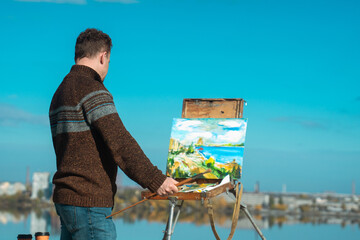 A man paints a vibrant landscape on an easel with a bright blue sky in the background, outdoors.