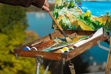 A close up of an artist's easel and palette outdoors with a vibrant landscape painting in progress.