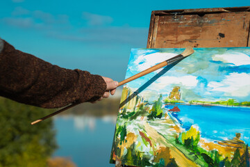A man paints a vibrant landscape on an easel with a bright blue sky in the background, outdoors.