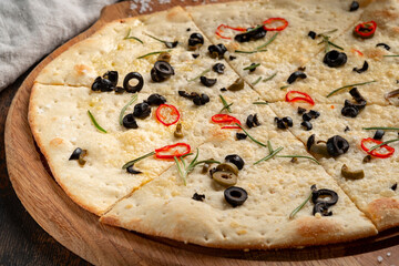 Homemade focaccia with black olives, red chili rings, rosemary and sea salt on wooden board. Authentic Italian flatbread, rustic bakery style, traditional recipe.