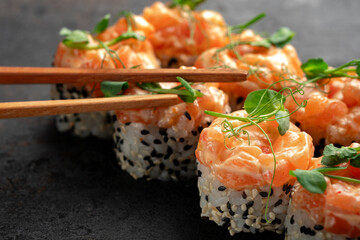 Elegant salmon sushi rolls crowned with creamy salmon tartare and fresh microgreens, served with wooden chopsticks on a dark textured background. Luxury Japanese cuisine, gourmet seafood roll.