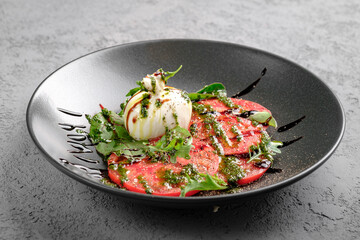 Fresh tomato carpaccio with creamy burrata, pesto drizzle, fresh basil leaves and balsamic glaze on black plate. Elegant Italian appetizer, gourmet salad-style dish, restaurant presentation, close up
