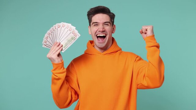 Happy young man wears orange hoodie hold in hand fan of cash money in dollar banknotes doing winner gesture celebrate isolated on plain pastel light blue background studio portrait. Lifestyle concept