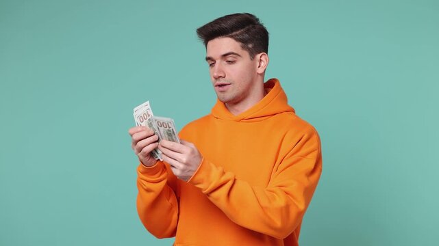 Happy fun accountant young man he wears orange hoodie hold in hand count stack of cash money in dollar banknotes isolated on plain pastel light blue cyan background studio portrait. Lifestyle concept