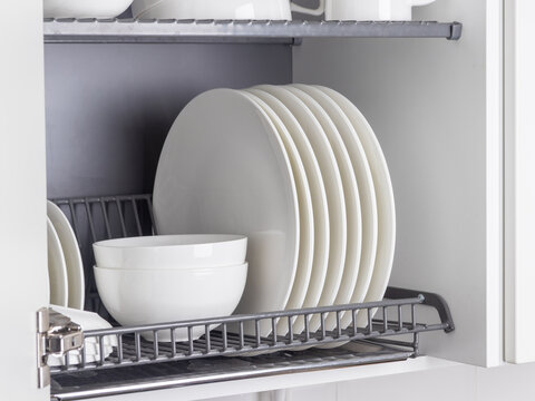A sleek metal dish-drying rack is neatly integrated into a wall cabinet in a modern kitchen. The rack holds white ceramic kitchenware, plates and mugs, all arranged orderly