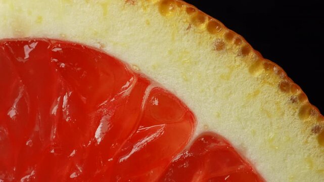 Macro close up of a red grapefruit slice slowly rotating, highlighting vibrant citrus flesh, rind texture, and glossy details against a black background. Ideal for food, color, and abstract concepts.