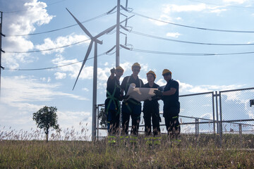 Workers hold plans near wind turbine at energy site in bright daylight surrounded by utility poles and tall grass
