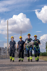 Group of construction workers walk down a path holding plans while wearing hard hats under a bright sky and fluffy clouds