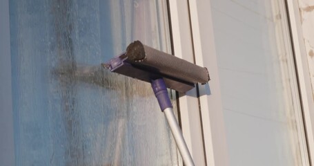 A hardworking woman cleans a dirty window pane at home with a professional brush on a telescopic pole. The woman cleans the windows and ensures flawless transparency in the sunlight.