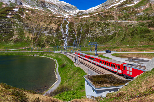 View of a local train traveling along the Lake Oberalp with snowy alpine mountains in background at Oberalp Pass, Andermatt, Switzerland