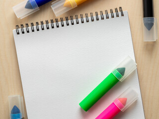 White spiral-bound notepad on a light wood surface with colorful washable crayons around it....