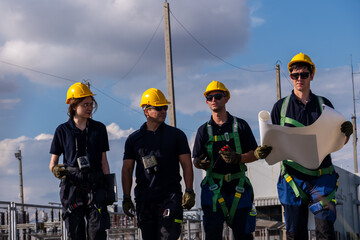 Workers walk on site at industrial location while holding plans and wearing safety gear during daylight hours
