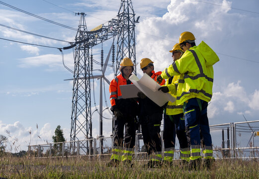 Workers discuss plans near electrical towers on a sunny day with blue skies and clouds in the background