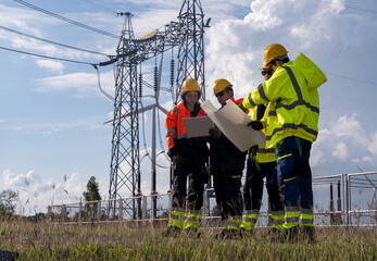 Group of workers examines plans at power station site with high voltage towers in the background on a sunny day