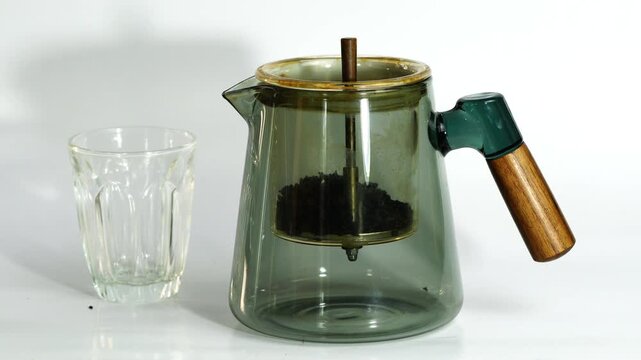 Making hot tea in an old transparent glass teapot on white background. Putting dried tea leaves into teapot, pouring boiling water and let the tea water flowing through strainer to the bollom of teapo