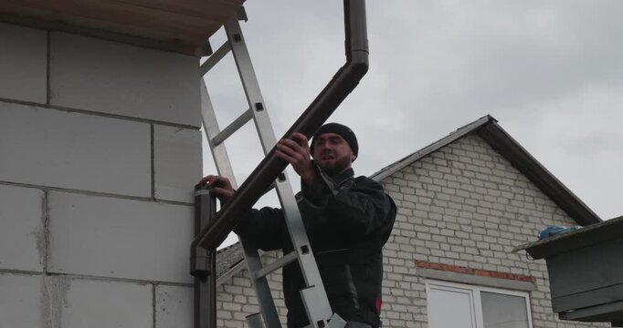 A Caucasian craftsman secures a gutter. A proud worker stands on a ladder, tightening a drainpipe joint against a cloudy sky. A man checks the conformity and strength of a brick house.