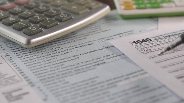 A person works on preparing 1040 tax forms in a home office setting. A calculator and various papers are spread out on a table, indicating the process of filing taxes.