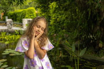A young girl stands beside a pond in a vibrant garden, wearing a floral dress. She rests her face...
