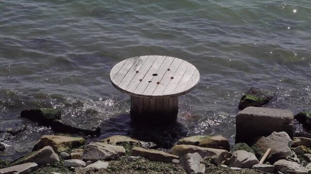 Large wooden cable spool lying in shallow water on the shore, representing environmental pollution, coastal waste and ecological issues affecting marine and shoreline areas