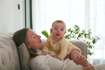 Young mom holding a joyful baby on the couch. Copy space, background.