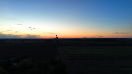 Fototapeta premium Cell tower standing amidst vast agricultural fields in the Po Valley, Italy, connecting the rural community under the warm hues of a vibrant sunset or twilight sky