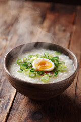 Bowl of congee with green onions and soy marinated egg in a ceramic bowl with rising steam on wooden table evoking gentle comfort and warmth  
