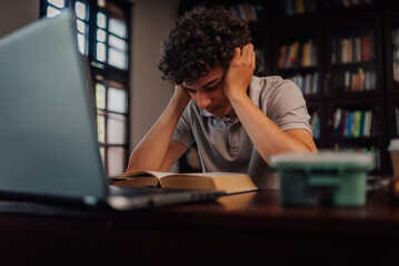 Tired student studying with book and laptop in library