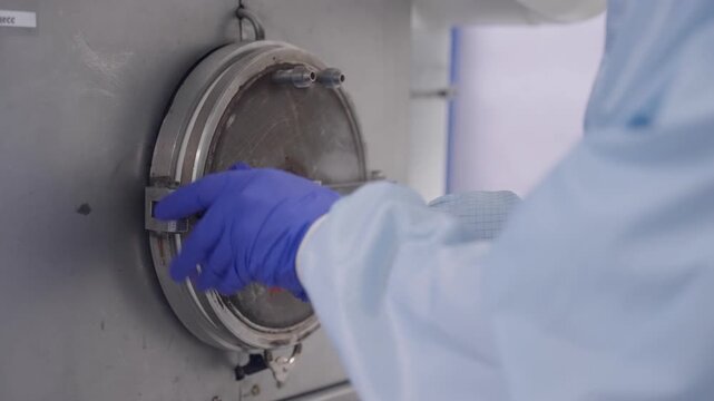 Gloved technician in sterile cleanroom operating a stainless steel vacuum chamber door, fastening clamps on sterilization or lyophilization equipment for controlled processing